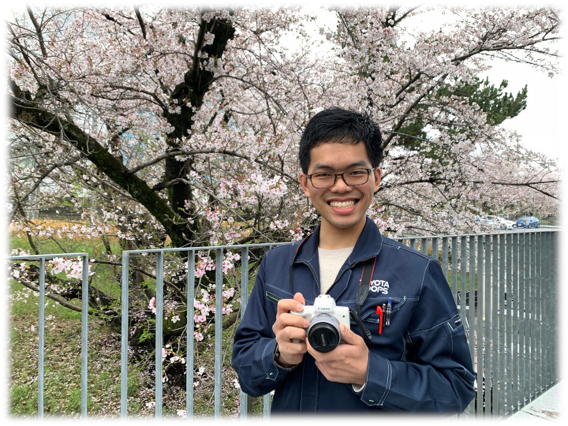 大澤宏輔さんの写真。桜の木をバックに、一眼レフカメラを構えている。