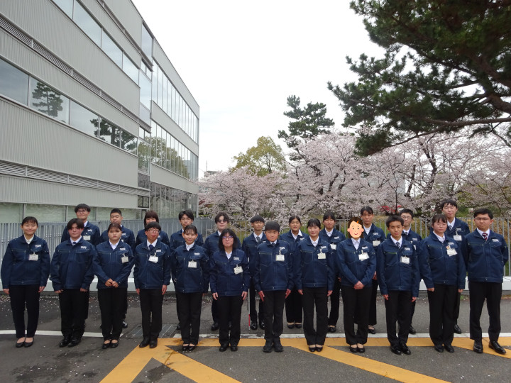 会社敷地内の駐車場にて撮られた集合写真。社屋と桜を背景に。