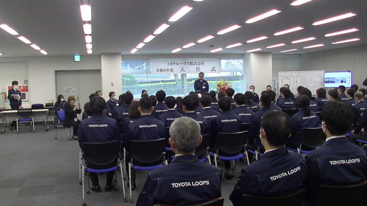 入社式の写真。広い空間の中に新入社員及び上長が集い、中央奥にて社長がお話をされている。