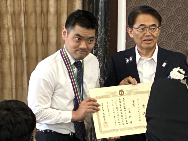 賞状を受け取った後、カメラに向けて賞状を見せている様子の写真。左手に山本選手、右手に大村知事。